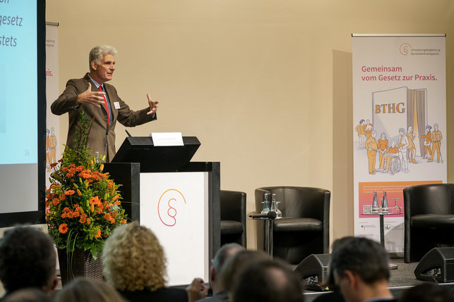 Das Bild zeigt Dr. Rolf Schmachtenberg, Leiter der Abteilung Teilhabe, Belange von Menschen mit Behinderungen, Soziale Entschädigung, Sozialhilfe im BMAS, auf der Bühne hinter dem Rednerpult.