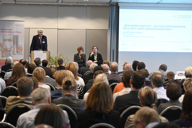 Das Foto zeigt den Veranstaltungsraum. Im Vordergrund ist das Publikum von hinten zu sehen. Im Hintergrund auf der Bühne steht Dr. Rolf Schmachtenberg, Staatssekretär im BMAS.