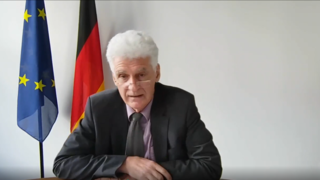 Screenshot von Dr. Rolf Schmachtenberg, Staatssekretär im BMAS, während seines Vortrags. Der Bildausschnitt zeigt ihn mit Gesicht und Oberkörper.