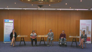 Das Foto zeigt fünf Menschen, die durch Tische getrennt nebeneinander sitzen. Der Moderator, der in der Mitte sitzt, unterhält sich mit Projektteilnehmerin Fiene Herkula, die links von ihm sitzt.