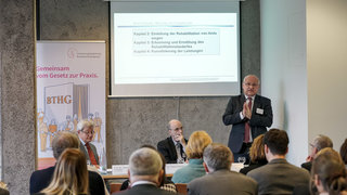 Das Bild zeigt von links nach rechts Herrn Prof. Dr. Johannes Schädler, Dr. Dieter Schartmann und Thomas Schmitt-Schäfer. Vor ihnen sitzen die Teilnehmenden des Forum 1. Hinter ihnen ist eine Leinwand, auf der die Präsentation zu sehen ist.