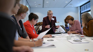 Das Bild zeigt eine Gruppe innerhalb einer Diskussionsstationen. Die Teilnehmenden sitzen am Tisch, notieren sich Dinge auf Moderationskarten und tauschen sich mit der Diskussionsleiterin aus.